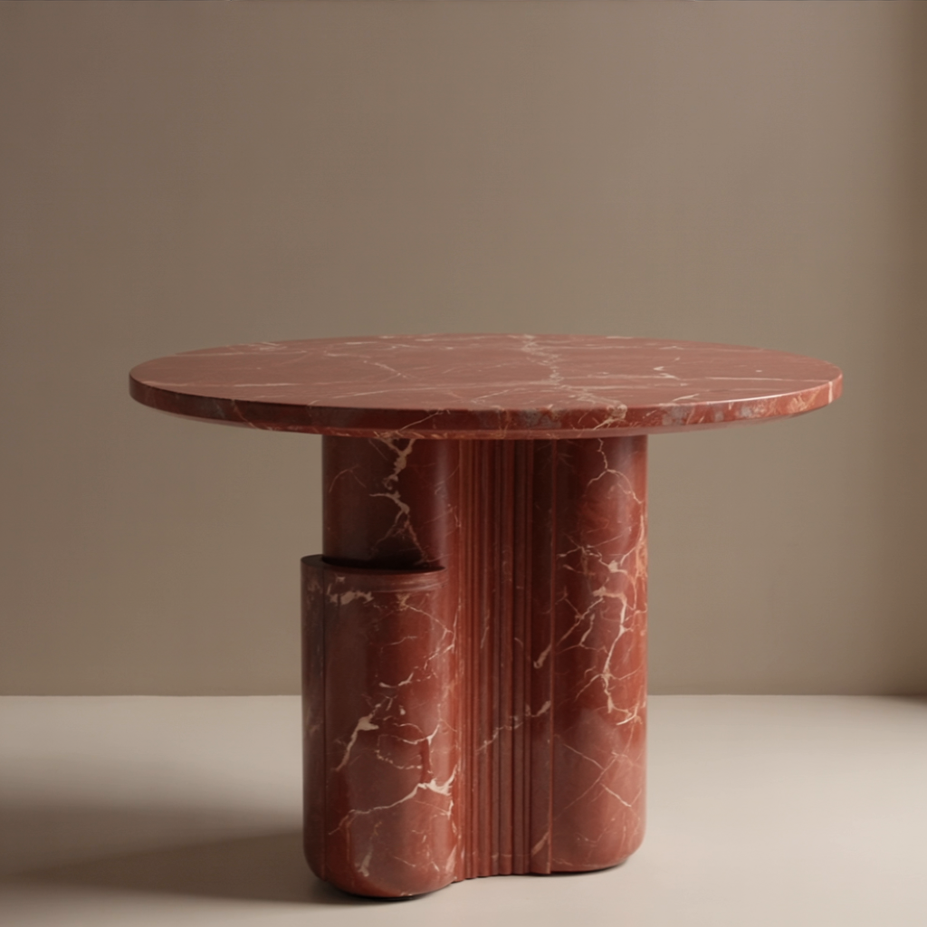 A round terracotta-red marble table with white and pink veining, its stepped pedestal base carrying a low semi-cylindrical shelf, against a sand-coloured studio wall